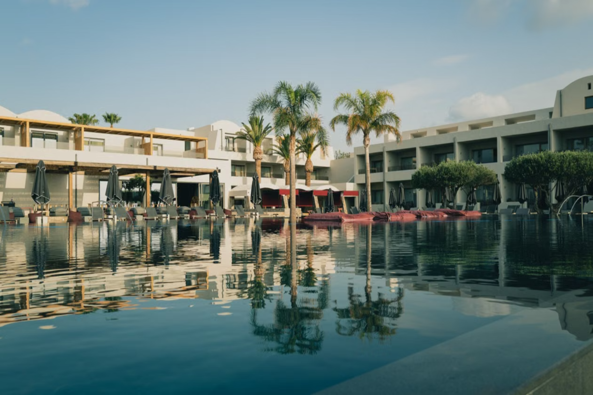 Lowrise resort in Greece with a large pool and palm trees under a clear blue sky.